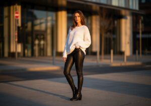Młoda kobieta w czarnych skórzanych legginsach i białym swetrze stoi na miejskiej ulicy podczas złotej godziny.
