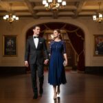 Para w eleganckich strojach wchodzi do klasycznego lobby teatru z czerwonymi zasłonami i drewnianą podłogą